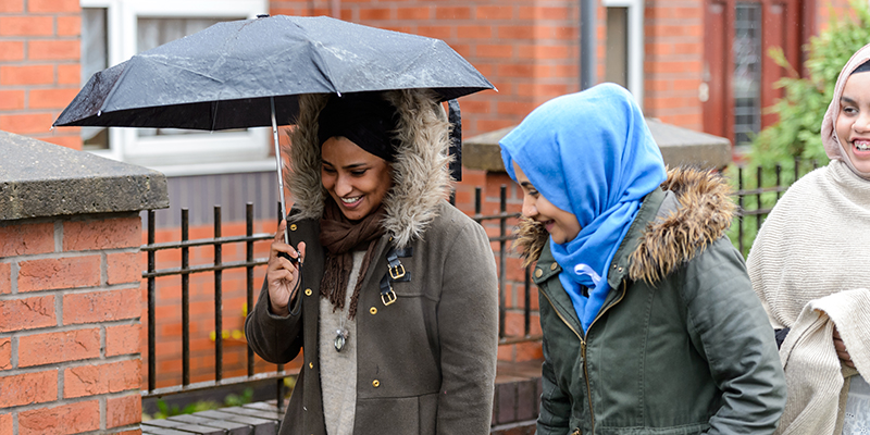A group of women walking along a street, one is holding an umbrella