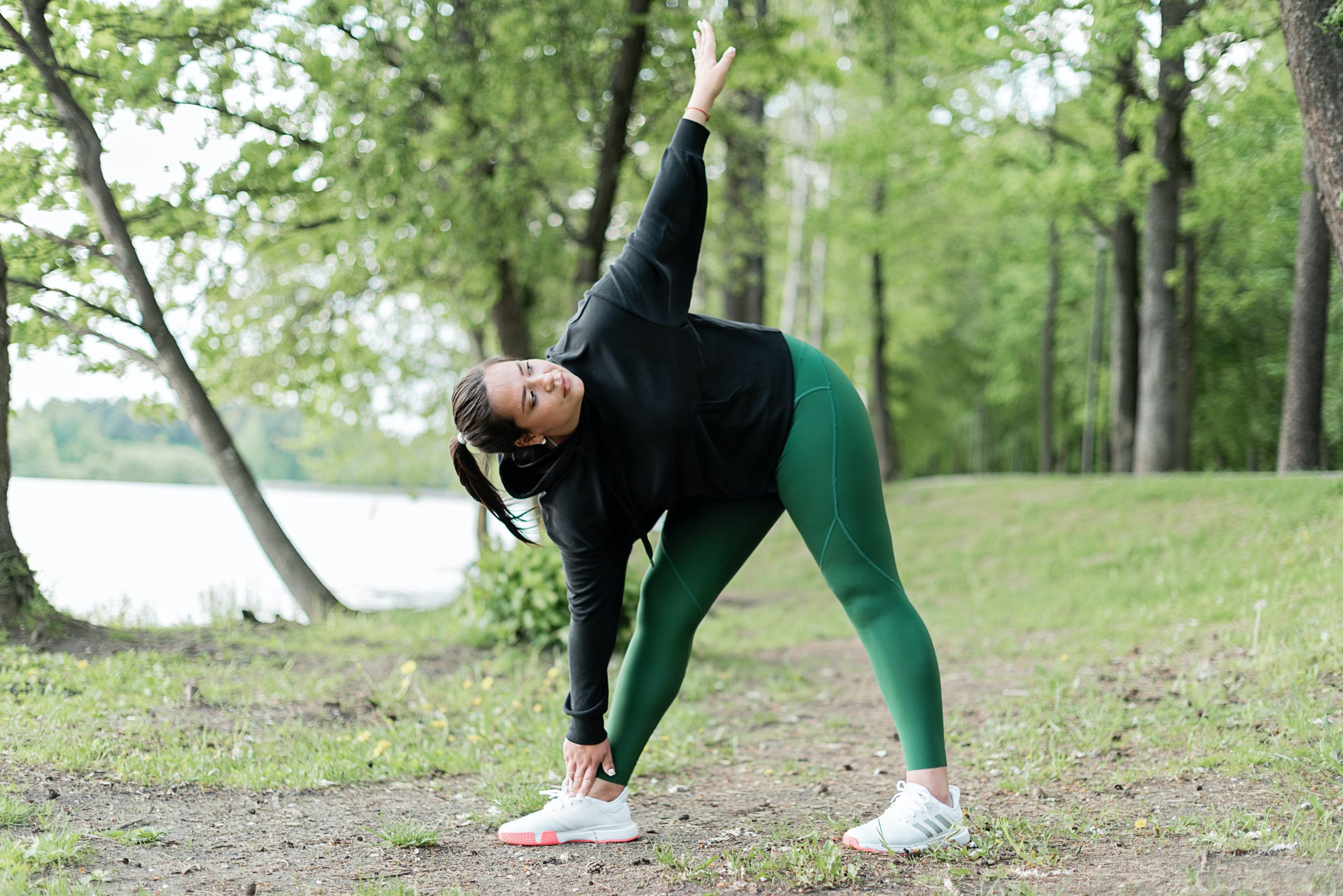 A woman stretching outside near a lake, touching her toe with one hand while the other reaches for the sky