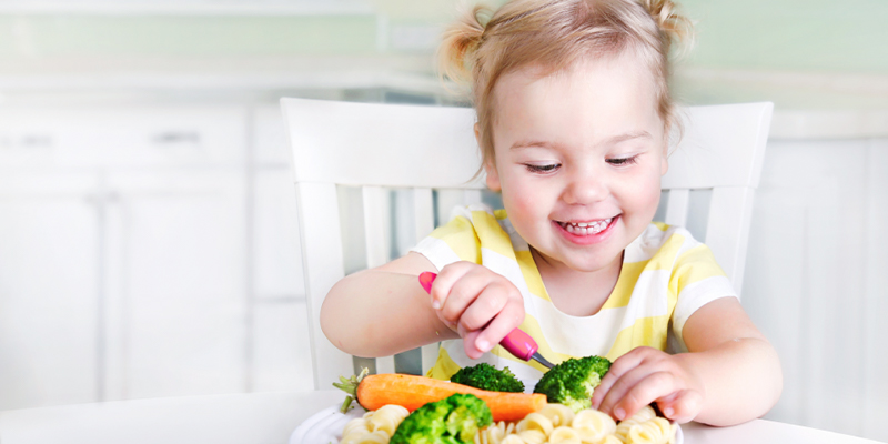 A young girl smiling as she eats a plate of pasta, broccoli and carrots