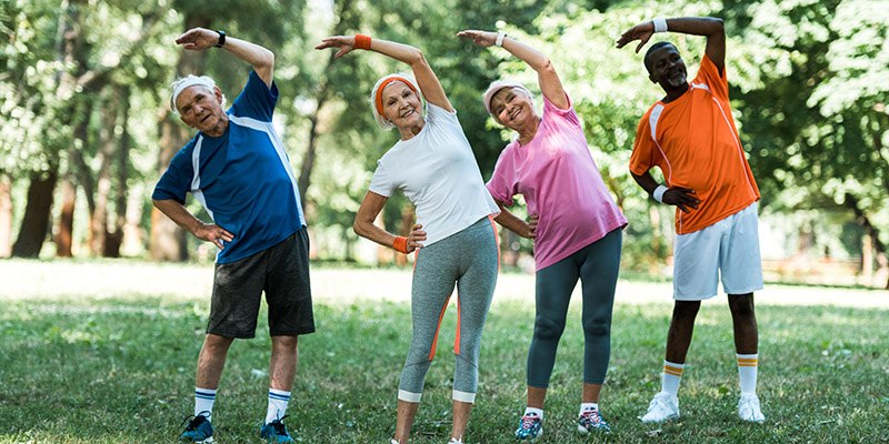 cheerful multicultural retired men and women standing with hands on hips while doing stretching exercise on grass