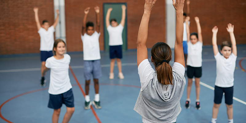 Rear view of coach and her students warming up during physical education in school gym.